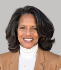 African American female wearing a taupe blazer with a white shirt and a delicate necklace. She has shoulder length dark hair with some gray strands
